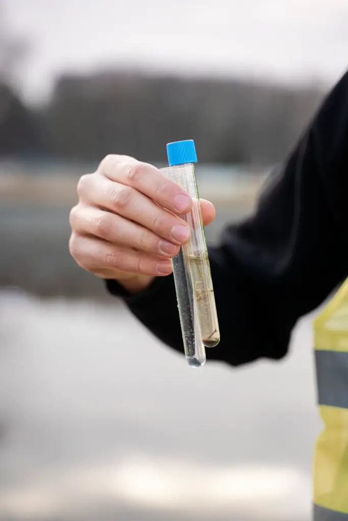 Hand holding two test tubes, one with clear water and one with murky water and sediment, near a body of water.